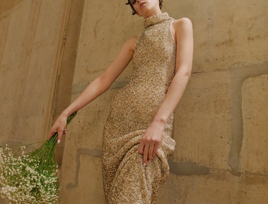 A model wearing a tan textured sleeveless turtleneck dress and holding a boquet of flowers. holding