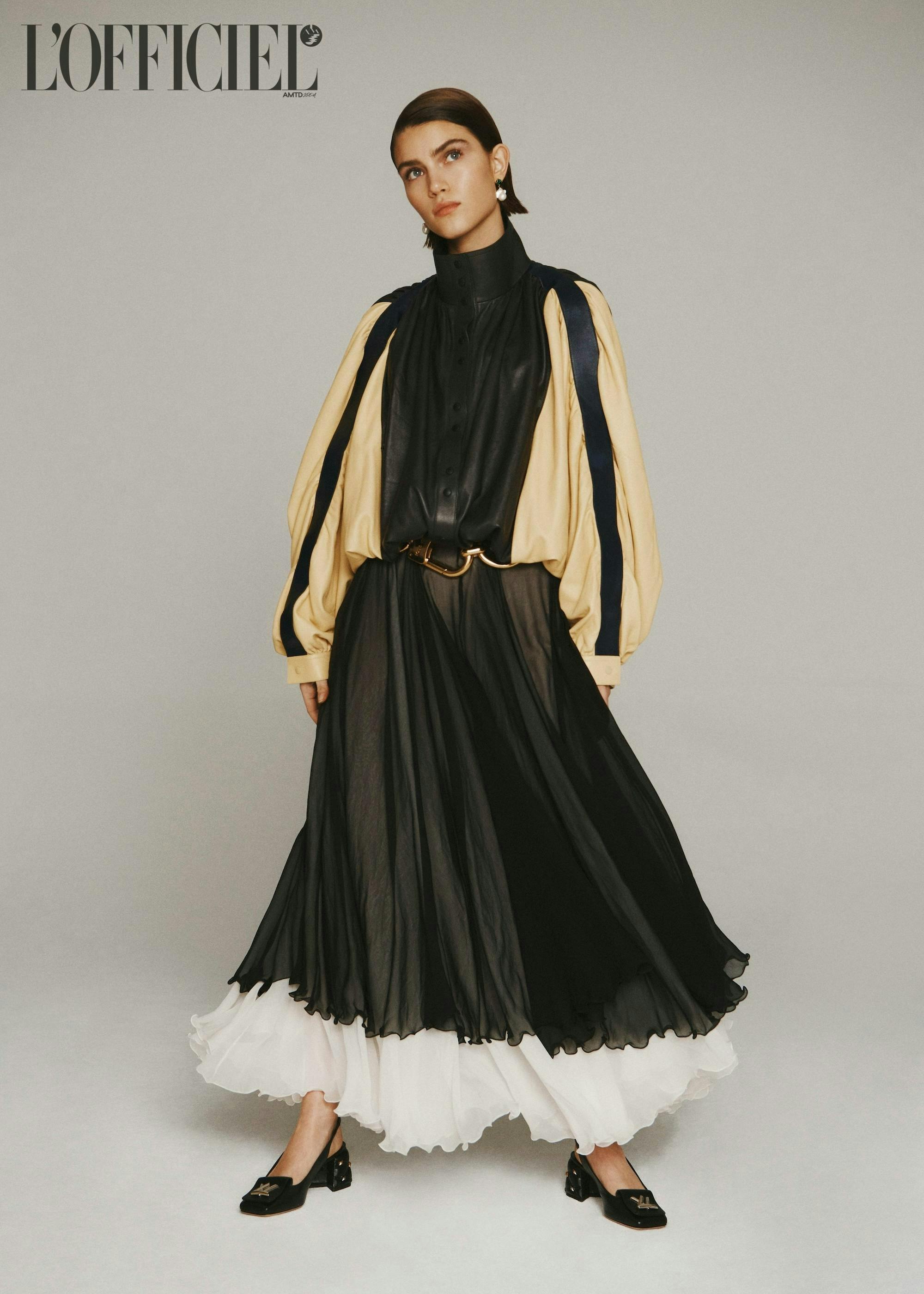 A model with a tan and black loose top, a long frilly black skirt with a white hem, black heels, a black and gold belt, and gold earrings.