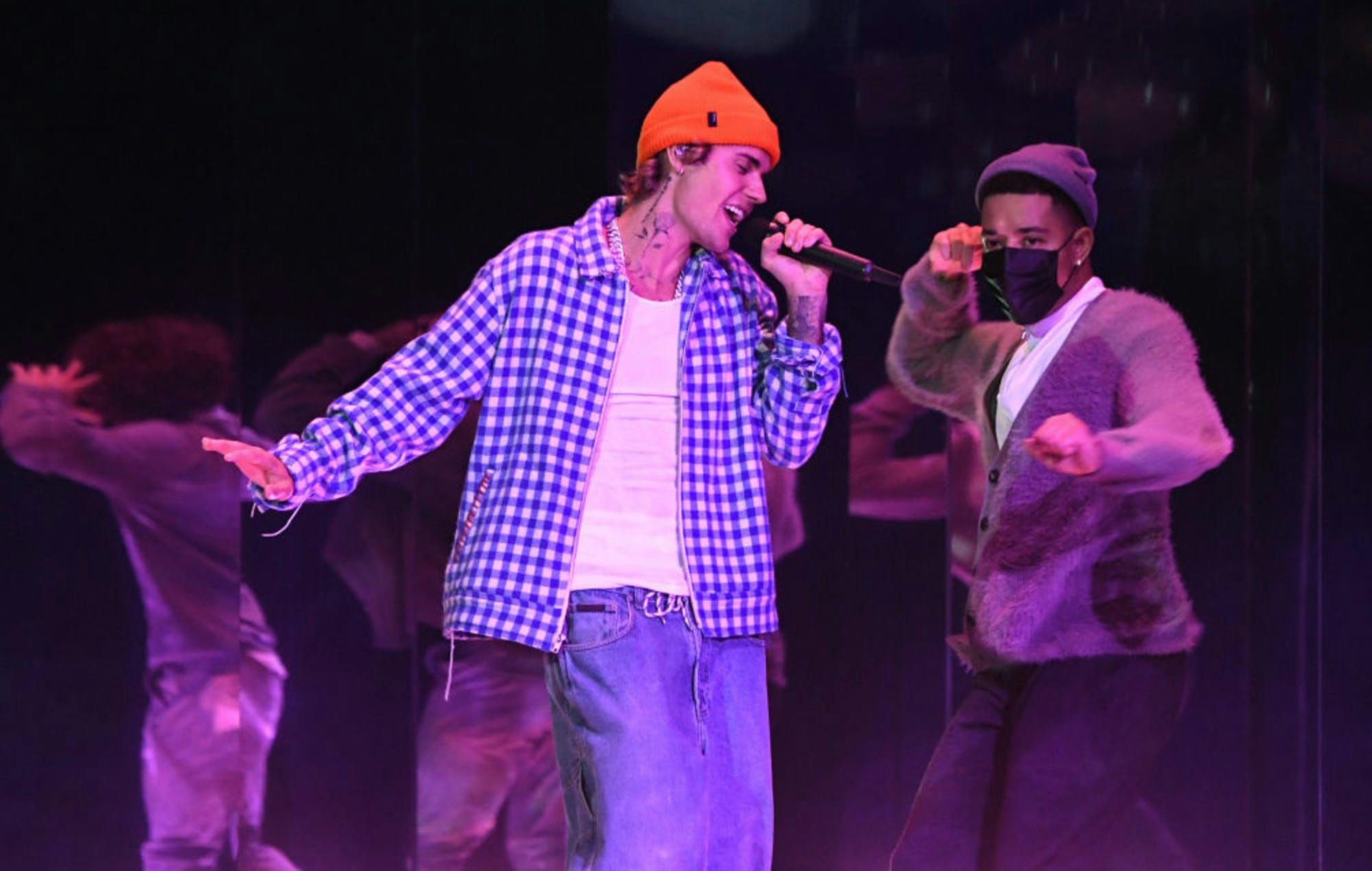 Justin Bieber singing while wearing jeans, a white shirt, a checkered jacket, and an orange beanie. Background dancers can be seen. 