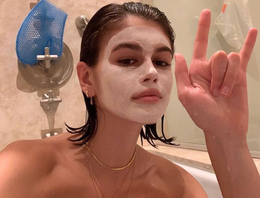 kaia gerber sitting in a bathtub with a white face mask on and hands up