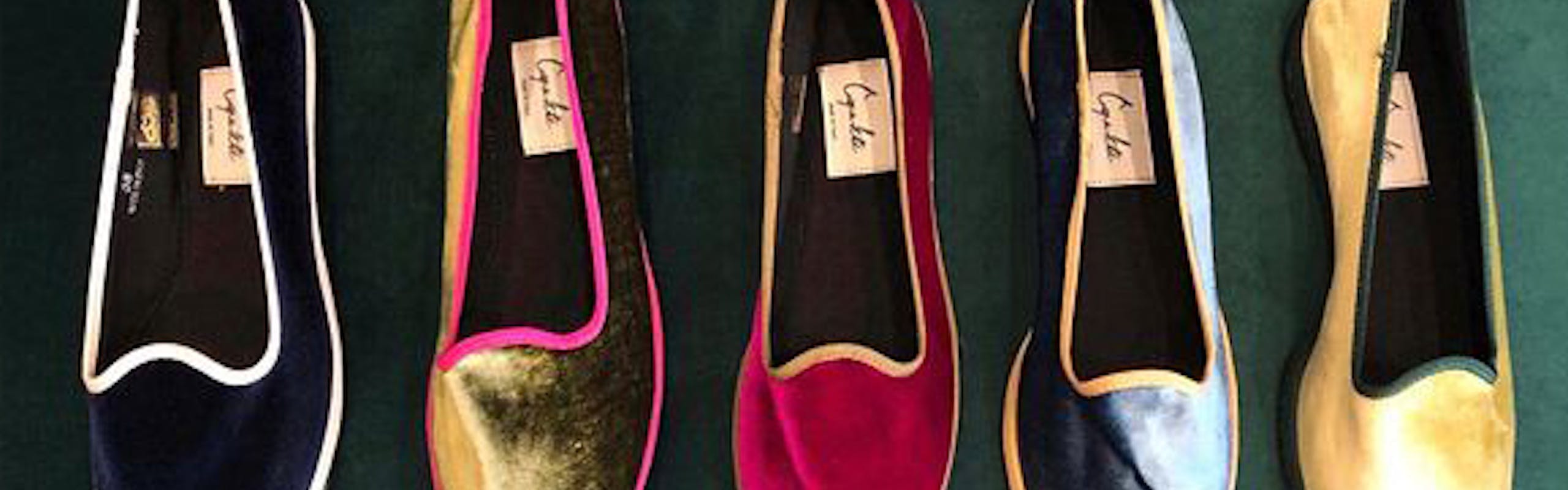 Five pairs of flat shoes are lined up side by side on a green velvet sofa. In order from left to the right the shoe colors are: black and white, gold and pink, red and pink, blue and orange, gold and green. 