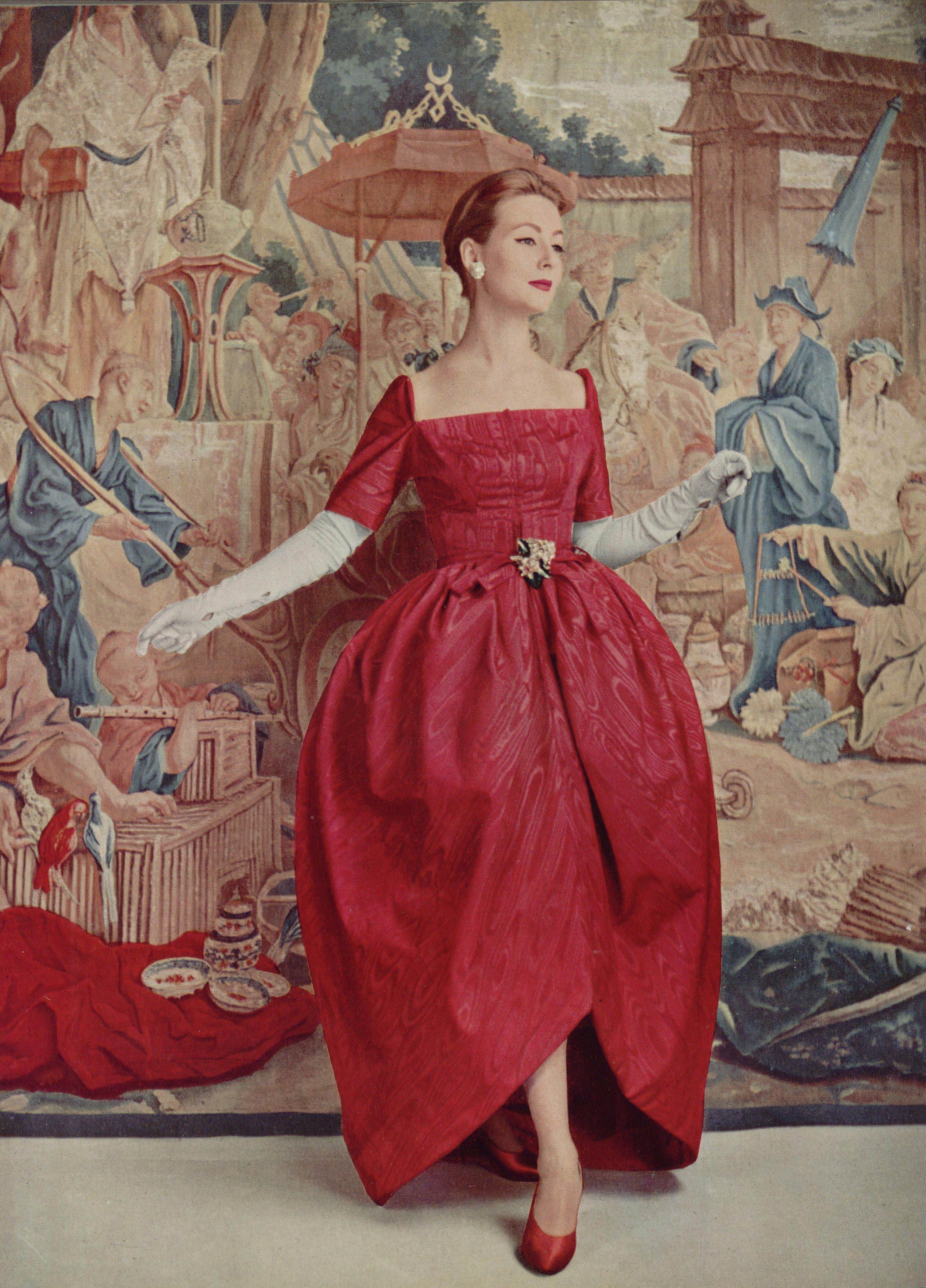 A model in a red dress with a square neckline, red heels and white gloves before a painted background.