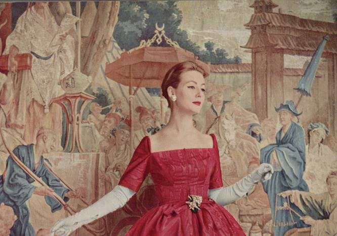 A model in a red dress with a square neckline, red heels and white gloves before a painted background.