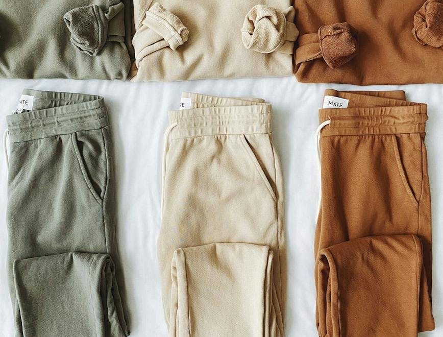 Three different sweatshirt and sweatpant sets folded on a bed. The set on the left is green, the set in the middle is beige, and the set on the right is dark orange in color. 