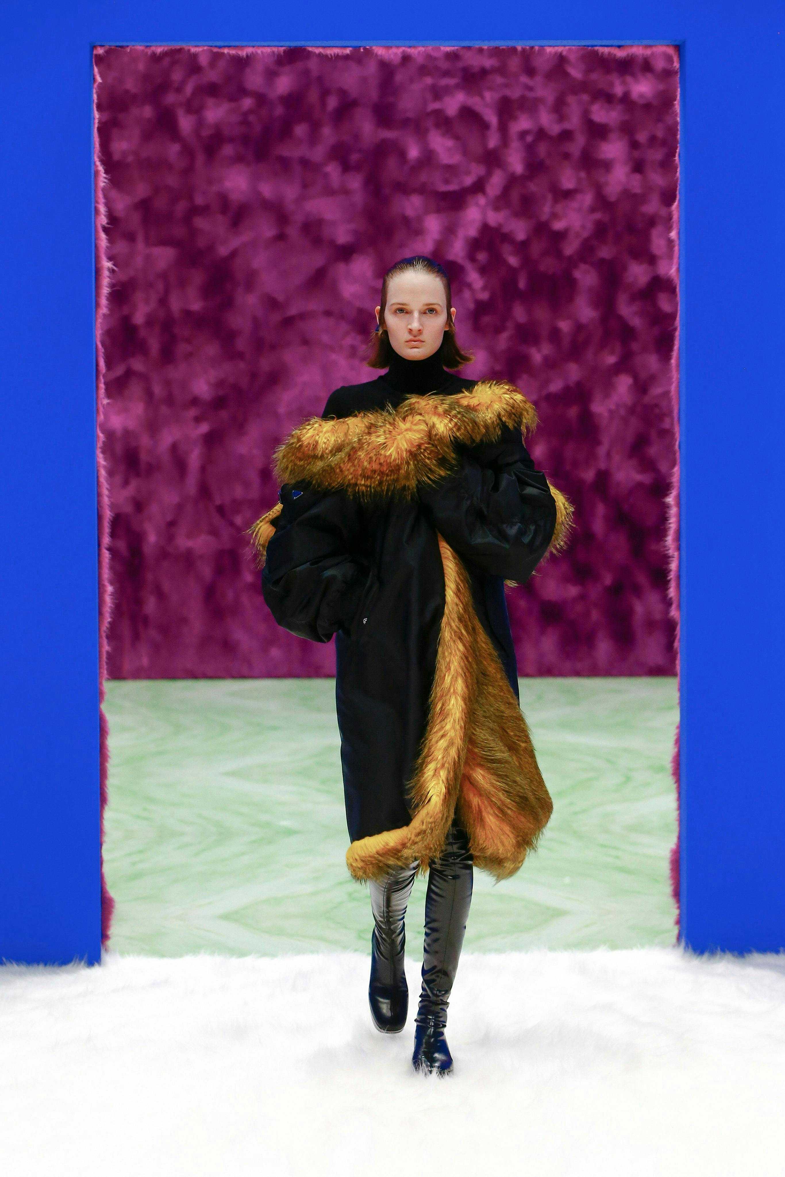 a model in the prada fall/winter 2021 show wears a faux fur coat and black boots and walks on a furry white carpet
