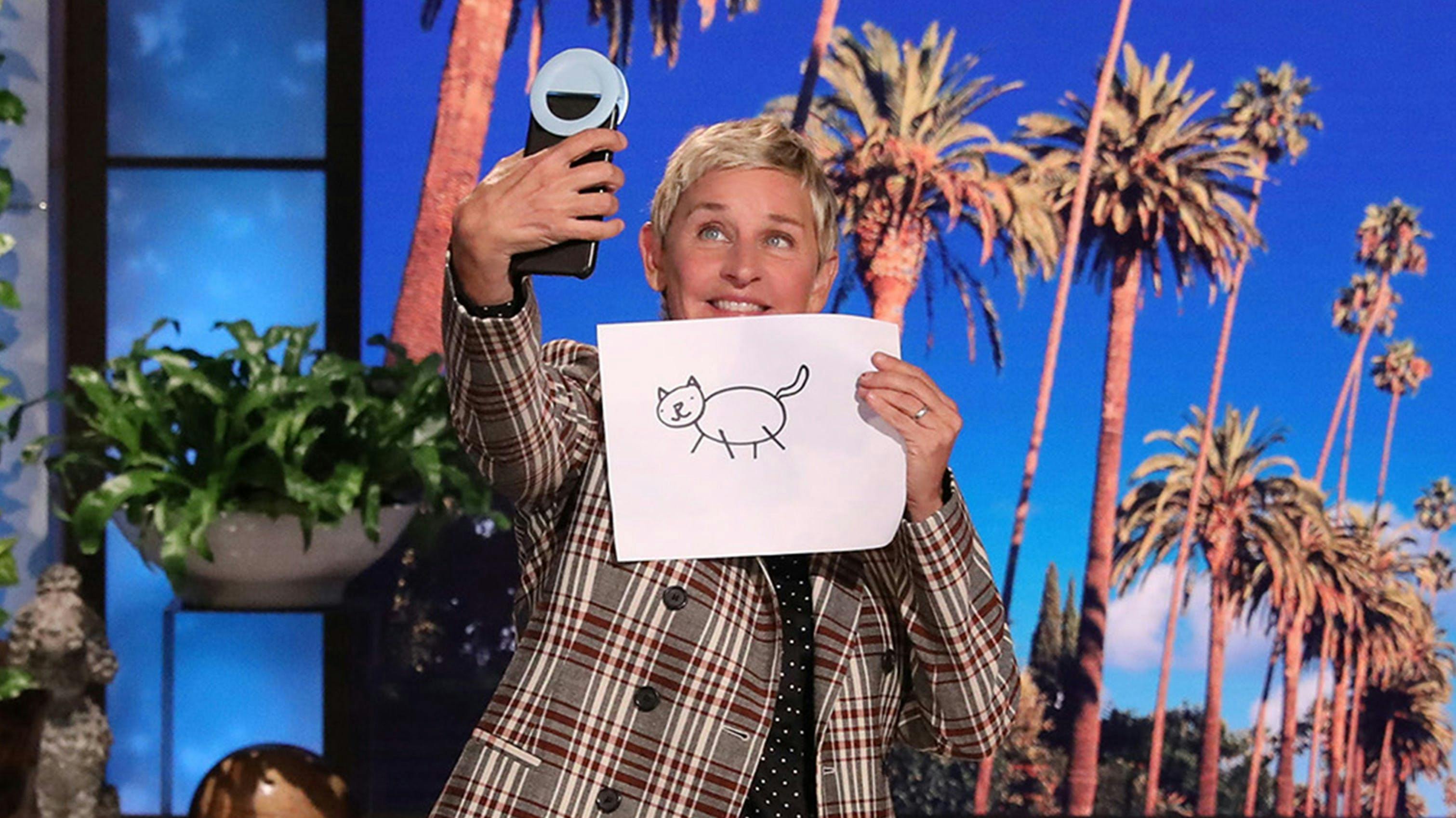 ellen degeneres taking a selfie with a drawing of a cartoon cat in front of a screen of palm trees