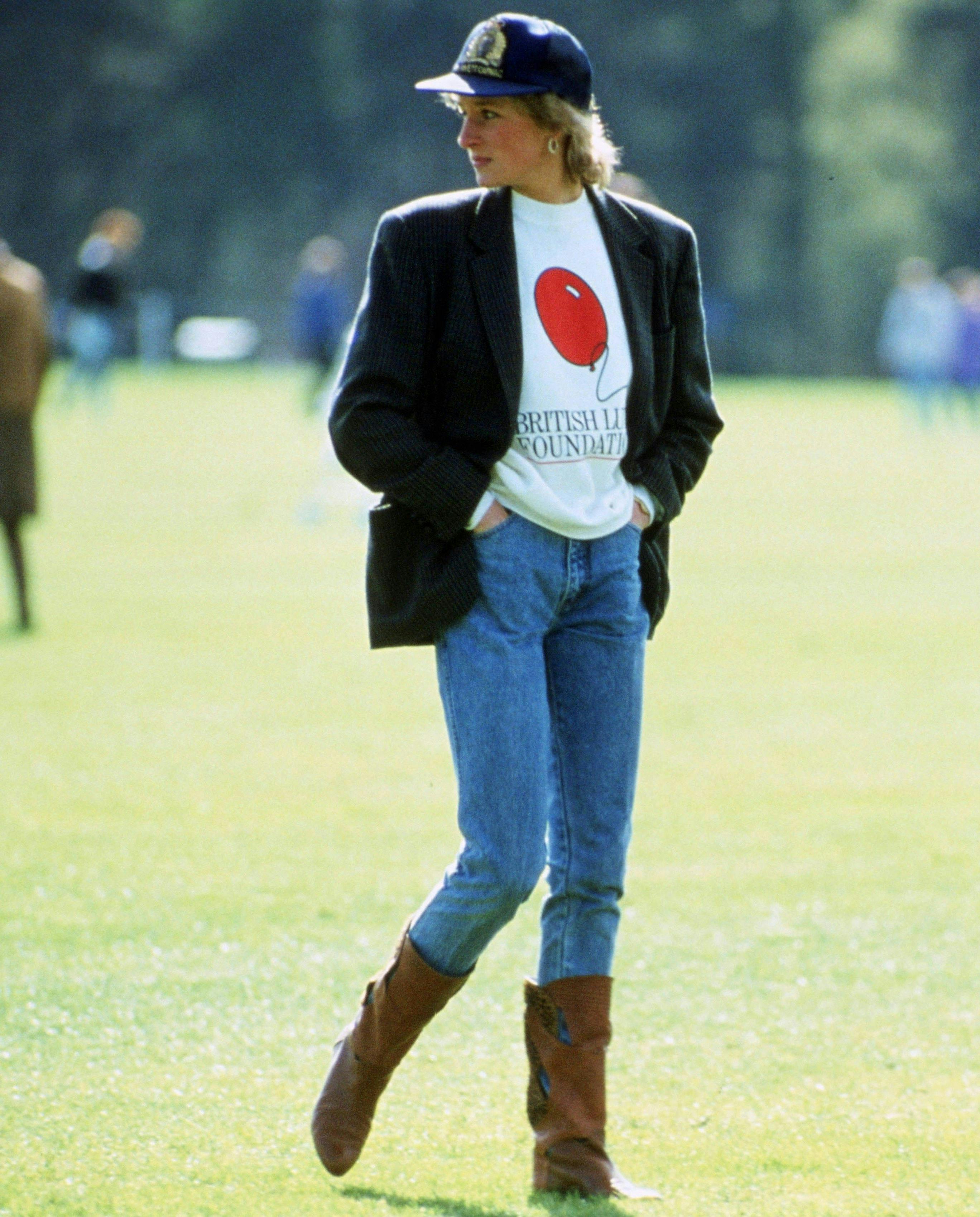 Princess Diana wears denim jeans, a white graphic sweatshirt, and a blazer thrown on top. She paired it with brown boots and a navy baseball cap. 