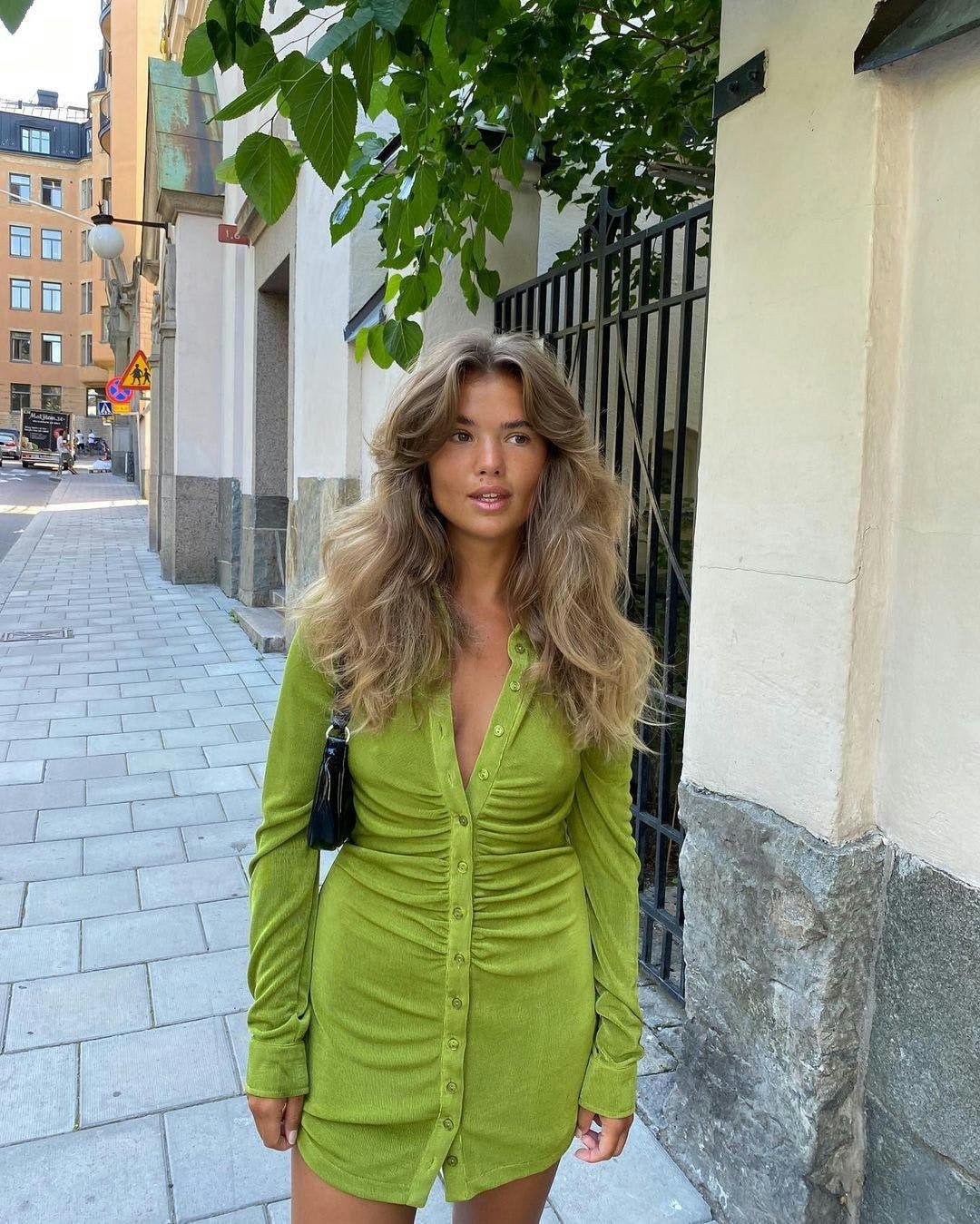 Fashion blogger with voluminous wavy hair wearing a green ruched mini dress.