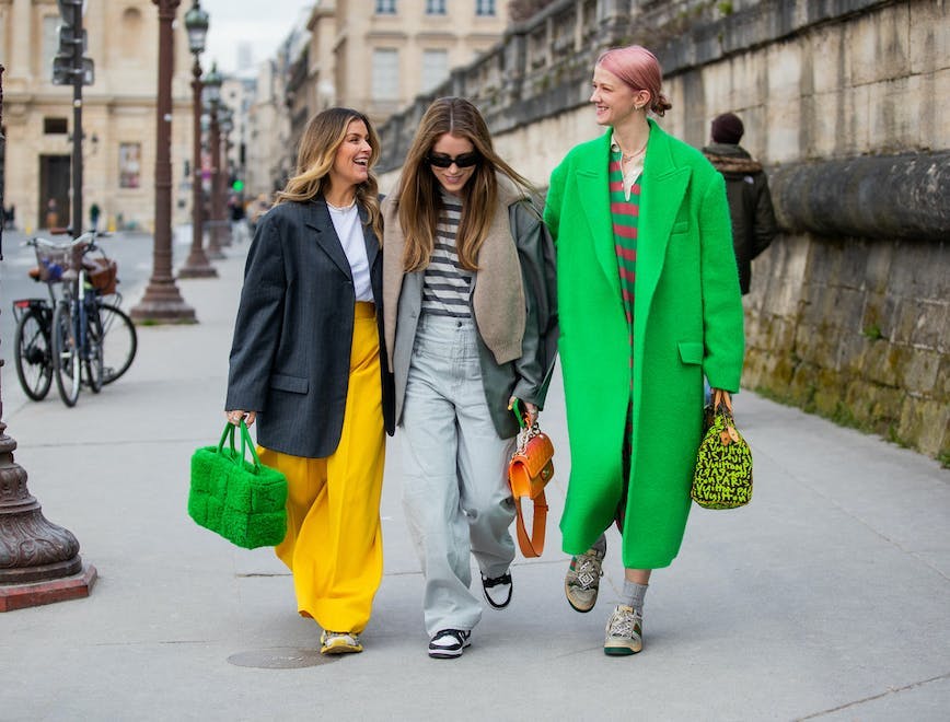 Three women walking down a street, left most woman wearong a black blazer with a white t-shirt and yellow pants, middle woman wearing a white and gray striped t shirt with a gray coat and light blue jeans, right most woman wearing a bright green over coat and a red and green striped shirt