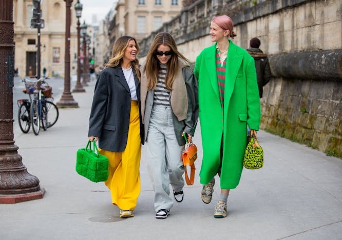 Three women walking down a street, left most woman wearong a black blazer with a white t-shirt and yellow pants, middle woman wearing a white and gray striped t shirt with a gray coat and light blue jeans, right most woman wearing a bright green over coat and a red and green striped shirt