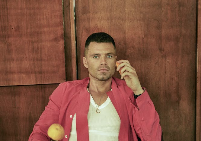Sebastian stan wears brown trousers, a black belt, a white tank top, and an open pink shirt. He sits on an arm chair with a wood panel behind him carrying an orange on a knife in one hand and an orange slice in the other.