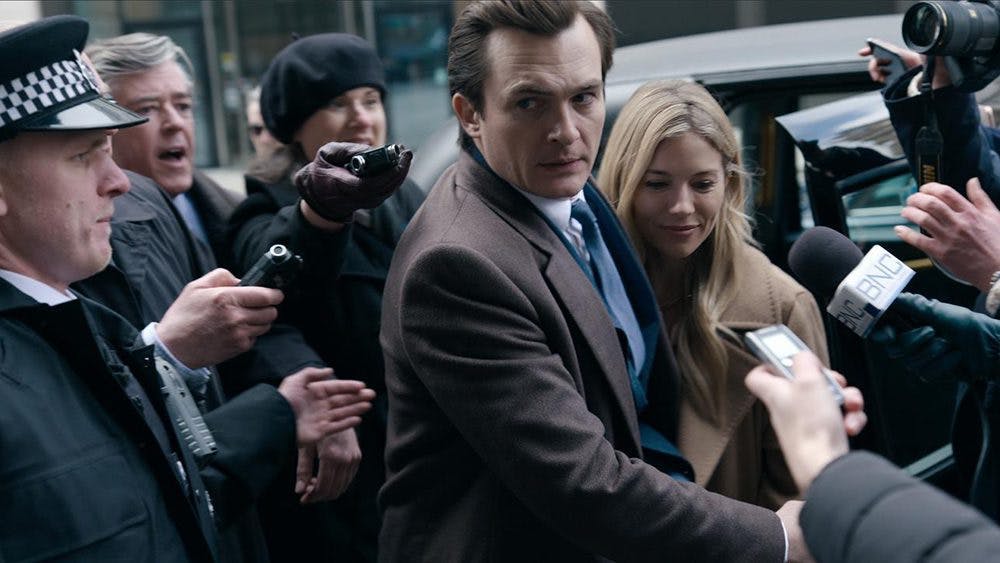 A shot from the new hulu show anatomy of a scandal. Police officers and reporters in the background outside and a man leading a woman in the foreground both in brown coats