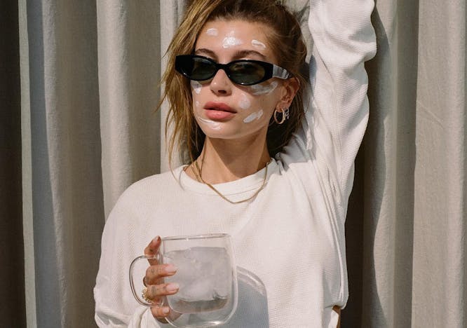 Hailey Baldwin in a cropped white sweatshirt and black sunglasses holding one arm up and holding a mug of ice water in the other. She is wearing her hair in a pony tail and has moisturizer place in lines around her face. 