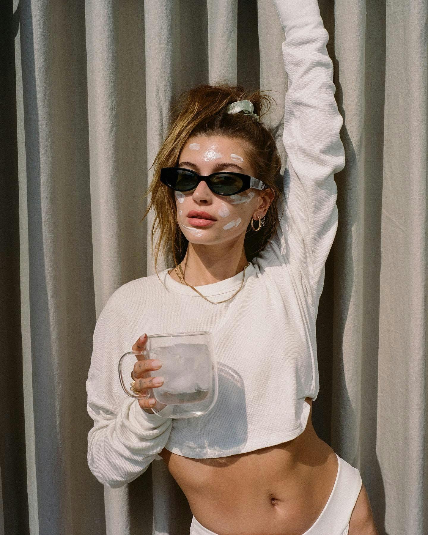 Hailey Baldwin in a cropped white sweatshirt and black sunglasses holding one arm up and holding a mug of ice water in the other. She is wearing her hair in a pony tail and has moisturizer place in lines around her face. 