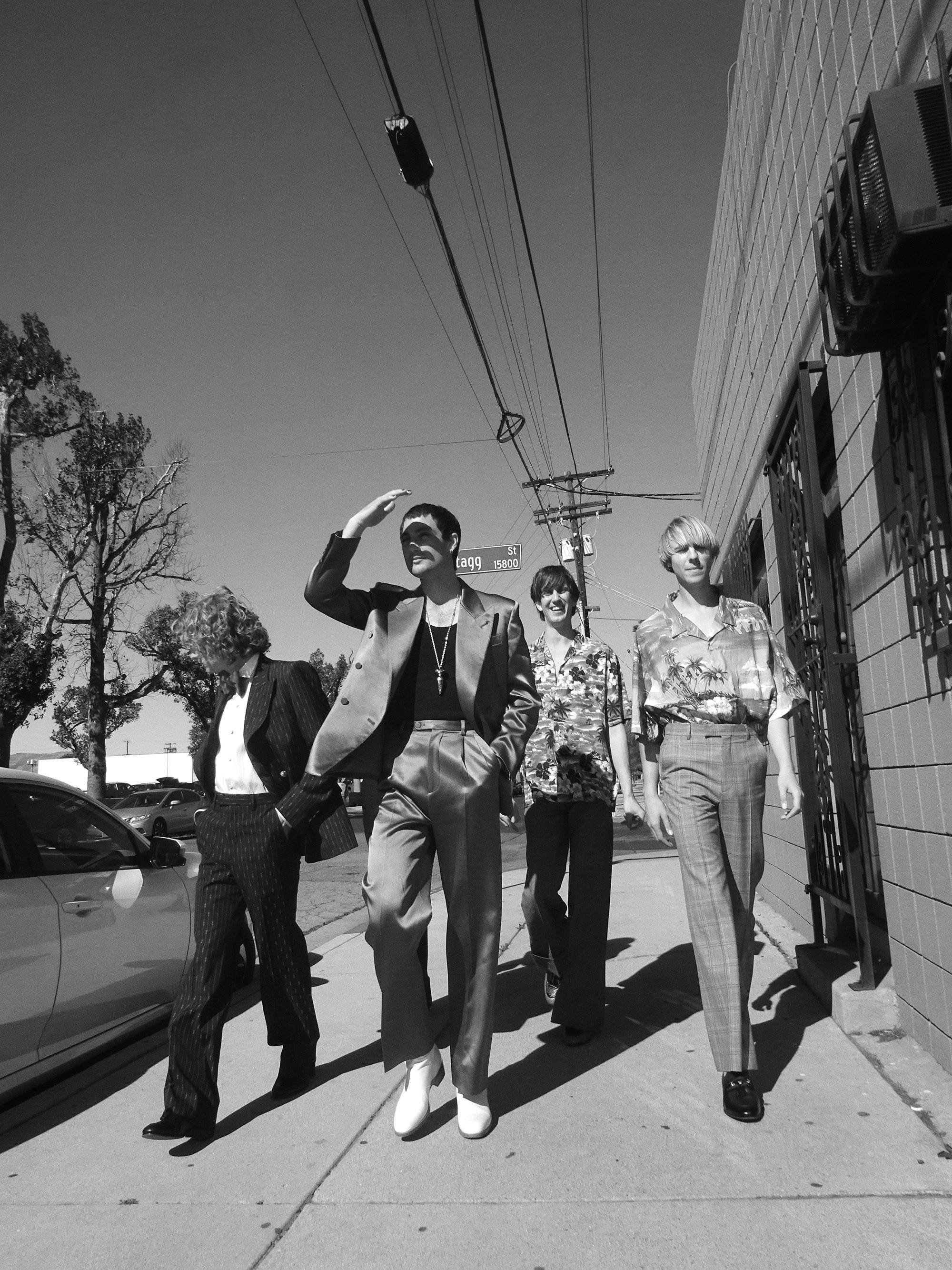 In a black and white photo, Australian band Parcels walk on the street beside a tree with a power line above them. 