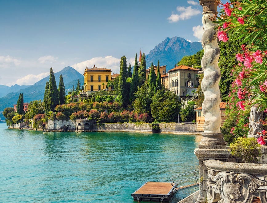 Lake Como Italy with italian houses, the blue lake, and mountains in the background. pink flowers in a bush on the side and a dock in the water with a staircase leading down beside it