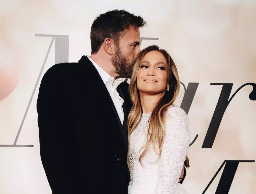 Ben Affleck kissing Jennifer Lopez's head on the red carpet in front of a white backdrop.