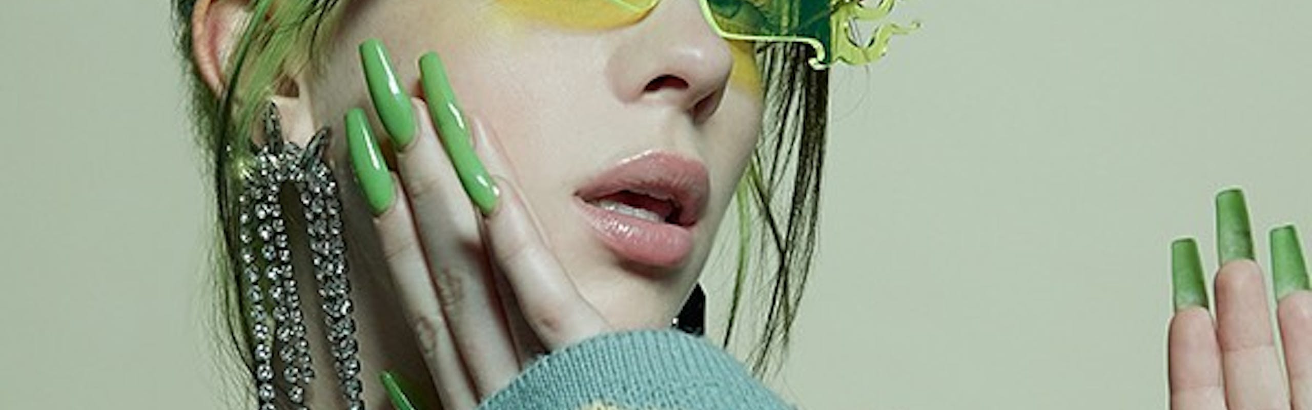 Billie Eilish with green and black hair and long green nails wearing a pair of green flame-shaped sunglasses