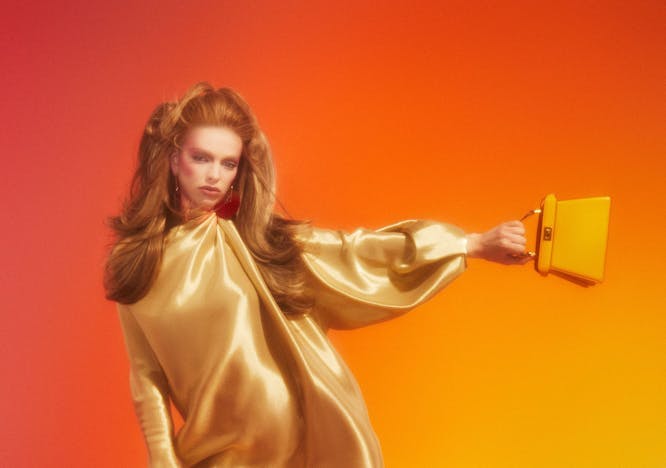 Model wears a gold dress and boots and holds a purse out to her side with an orange background
