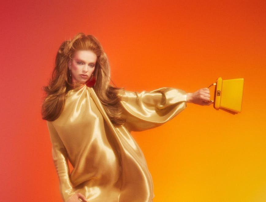 Model wears a gold dress and boots and holds a purse out to her side with an orange background