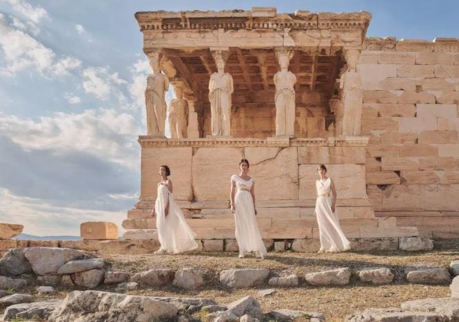 Three models in white dresses stand on rocks in front of Greek ruins in Athens