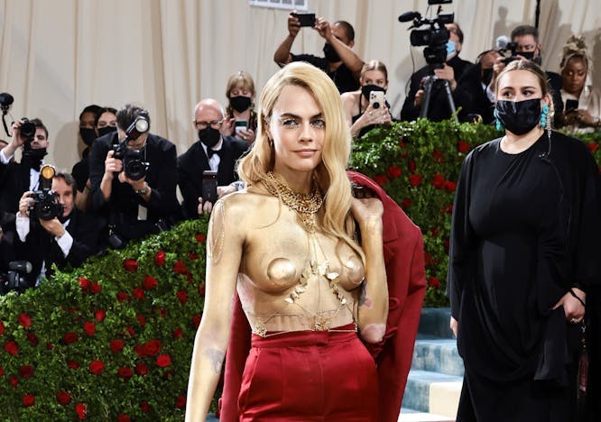 Cara Delevingne at the 2022 Met Gala with her body painted gold, wearing gold pasties and body chain, a red suit and a cane.