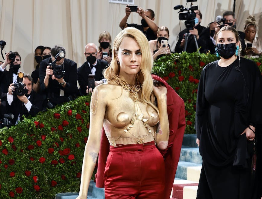 Cara Delevingne at the 2022 Met Gala with her body painted gold, wearing gold pasties and body chain, a red suit and a cane.