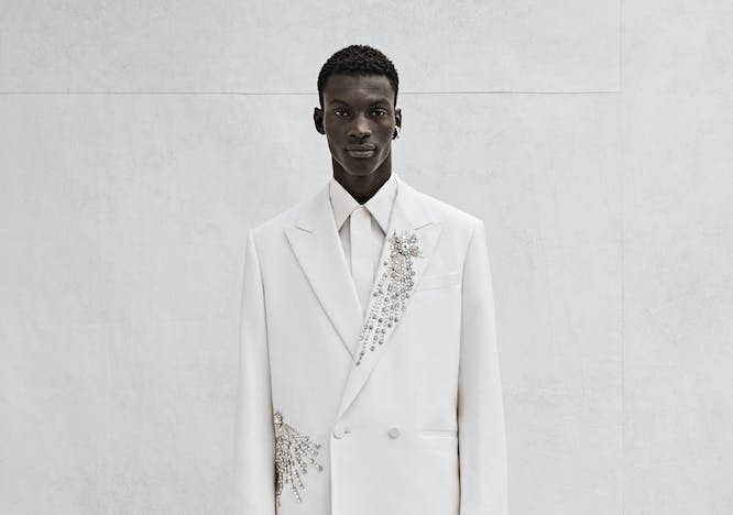A model poses in an all-white outfit. The suit jacket and pants have crystal embroidery. 