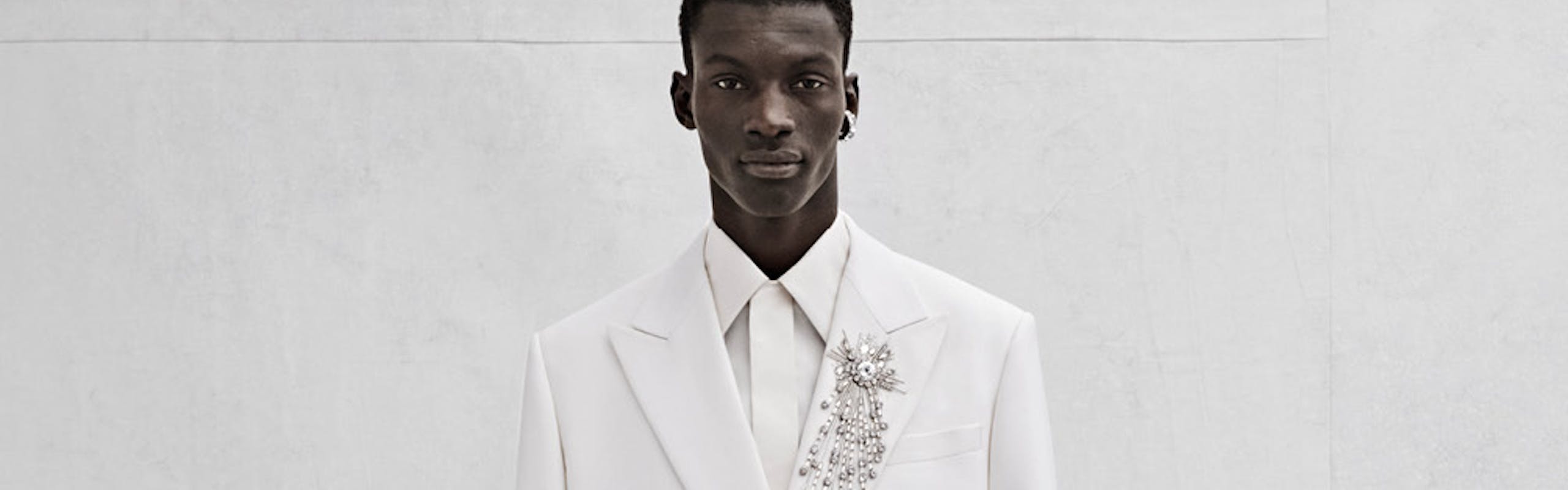 A model poses in an all-white outfit. The suit jacket and pants have crystal embroidery. 