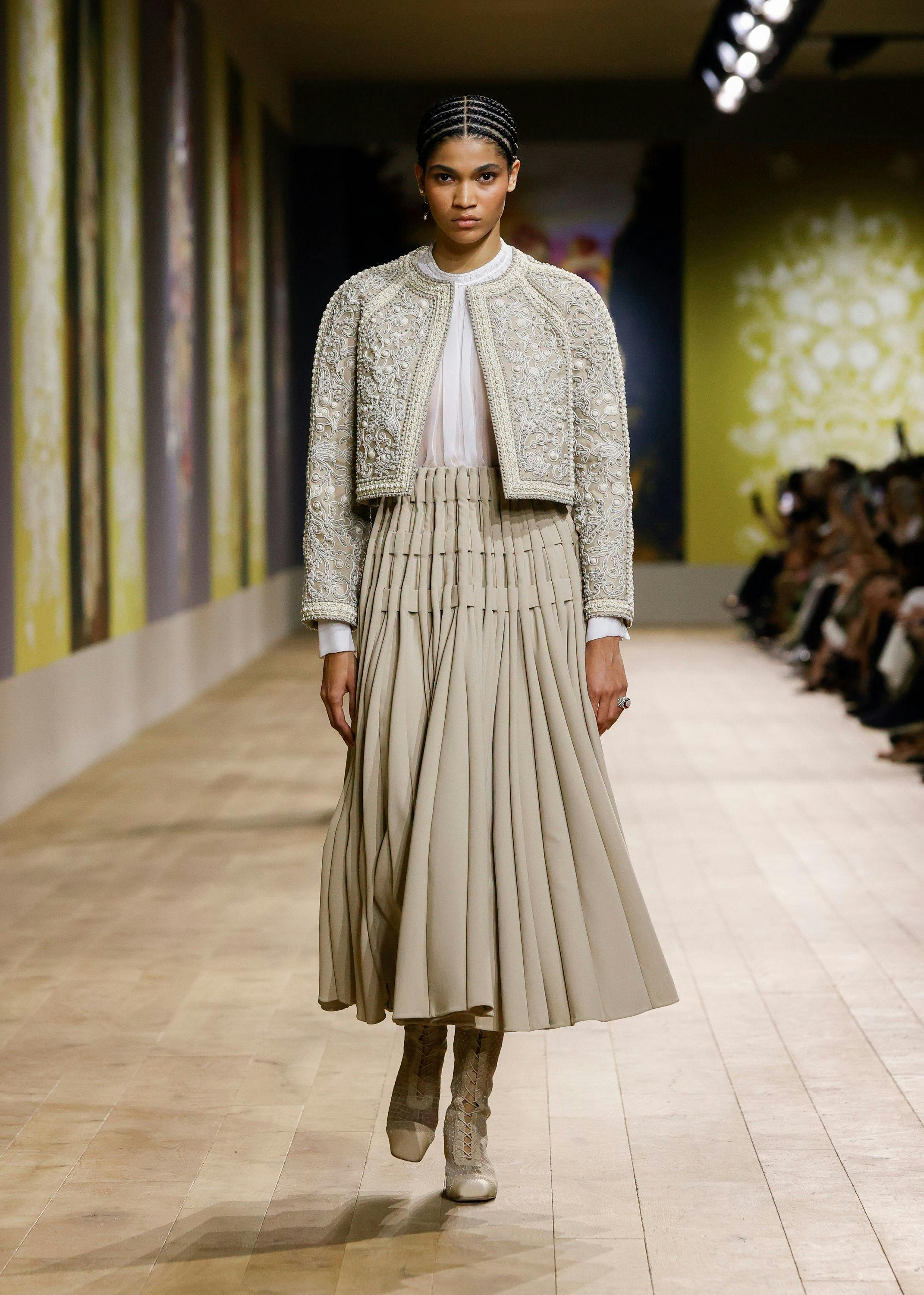 Model walking the Dior runway wearing an embroidered jacket and tan pleated skirt.