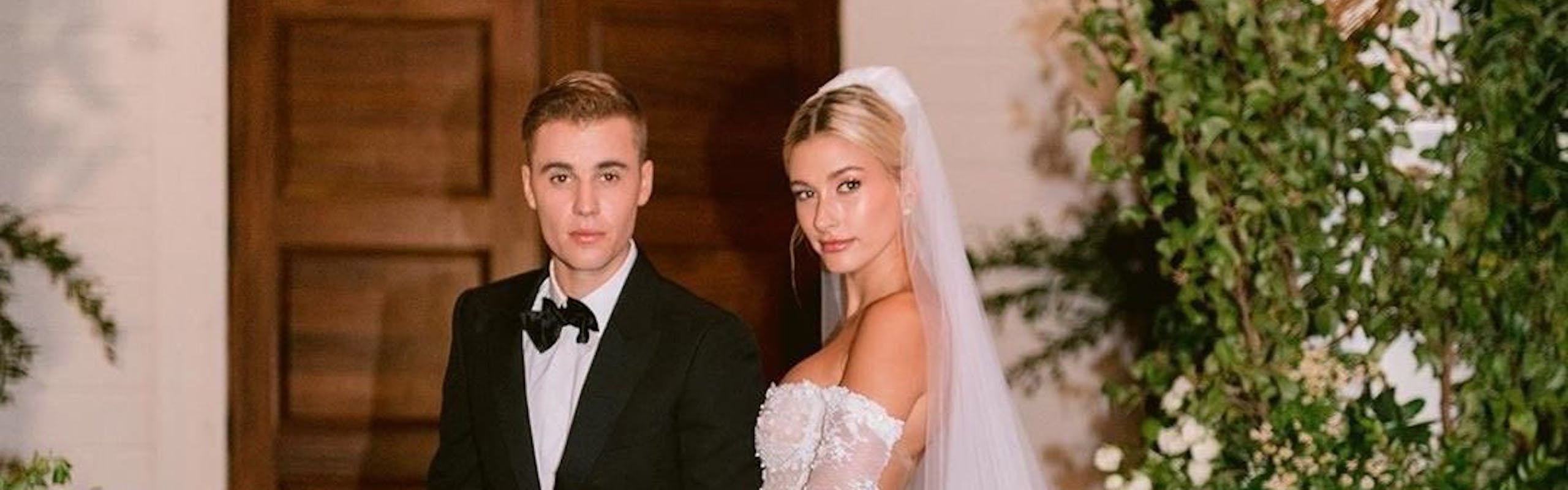 Justin and Hailey Bieber on their wedding day, with Justin wearing a tuxedo and Hailey wearing a white embroidered wedding gown on brick steps