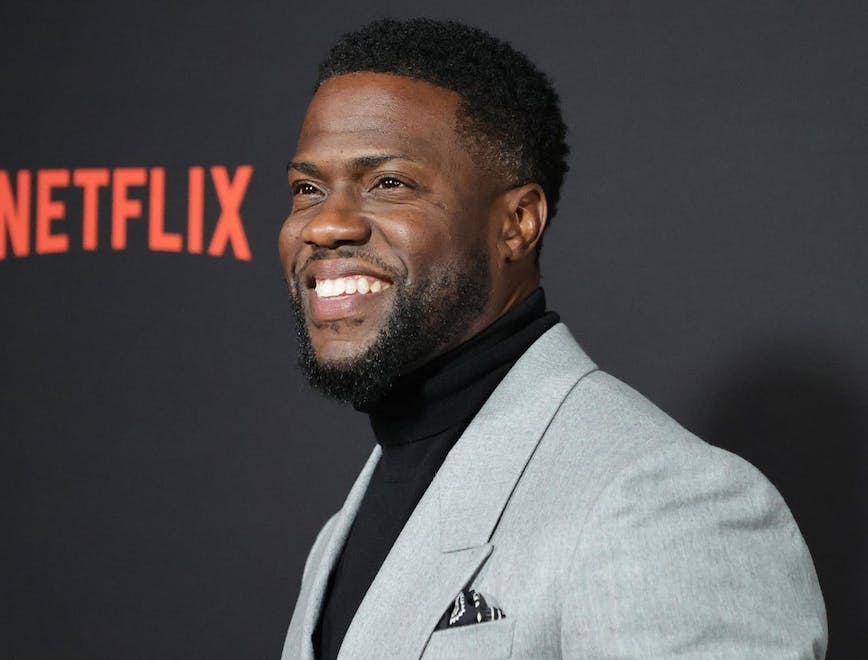 Closeup of Kevin Hart on a red carpet wearing a grey suit and black turtleneck. 