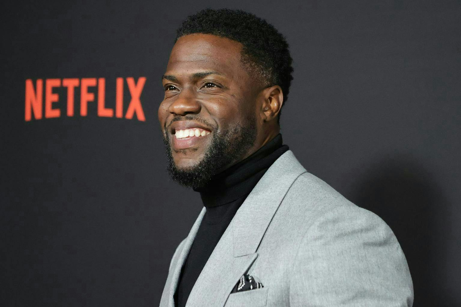 Closeup of Kevin Hart on a red carpet wearing a grey suit and black turtleneck. 