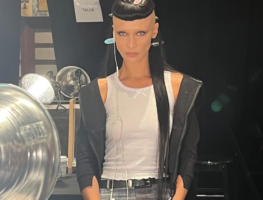 Bella Hadid backstage at Marc Jacobs with bleached eyebrows and a mullet. She is wearing a white tank top, black jacket, two-tone denim jeans and wired headphones.