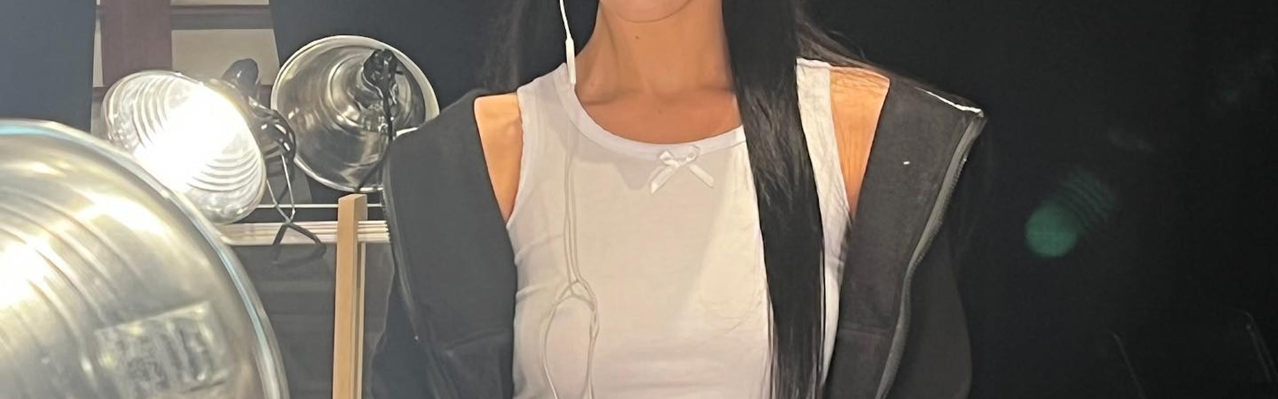 Bella Hadid backstage at Marc Jacobs with bleached eyebrows and a mullet. She is wearing a white tank top, black jacket, two-tone denim jeans and wired headphones.