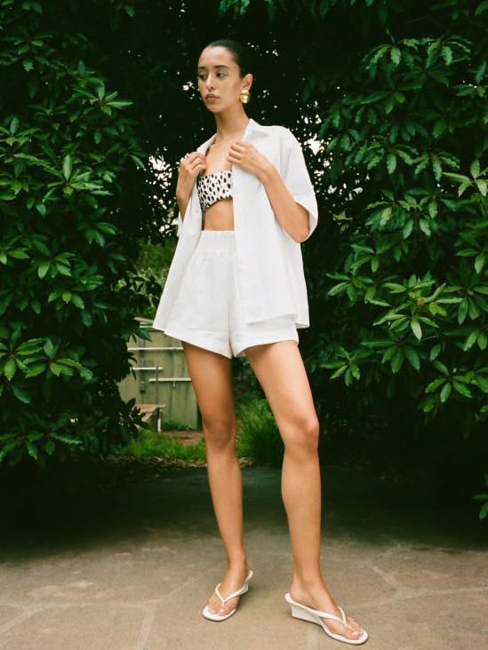 Photo of a model standing in a garden wearing a white linen button down and matching shorts with heeled flip flops and a bikini top.