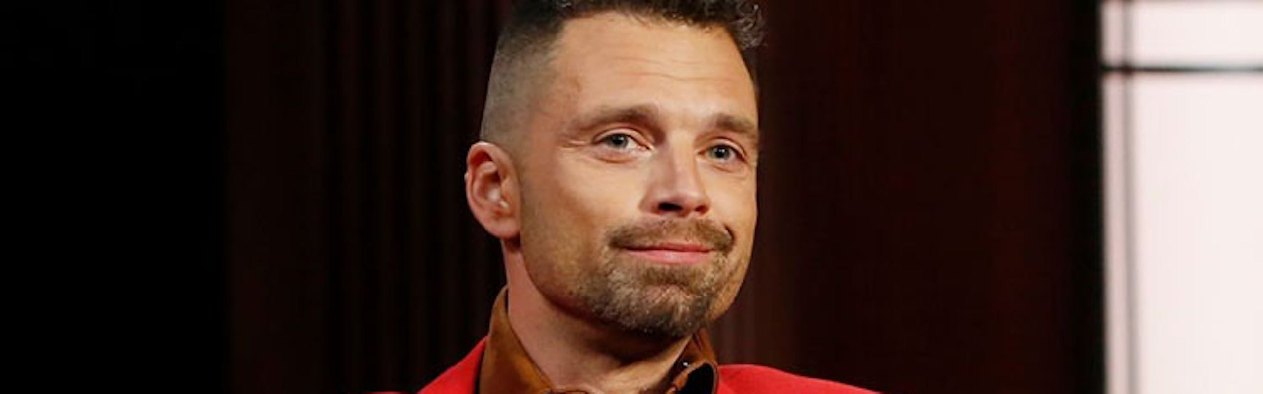 Sebastian stan in a red matching suit with a brown leather shirt underneath sitting with his hands crossed on a grey chair in front of a black wall