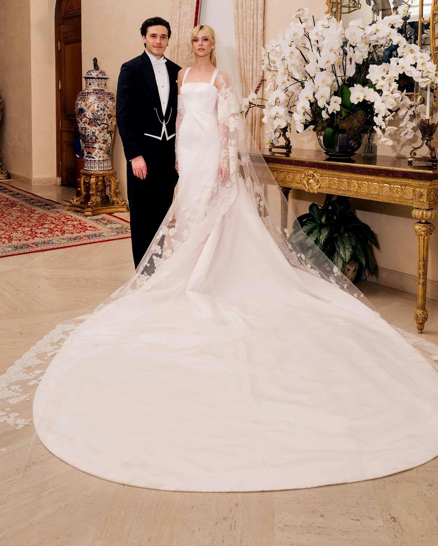Nicola Peltz in her white gown and cascading veil and Brooklyn Beckham in his black tuxedo posing after their wedding ceremony with Nicola's dress train pooled in front..