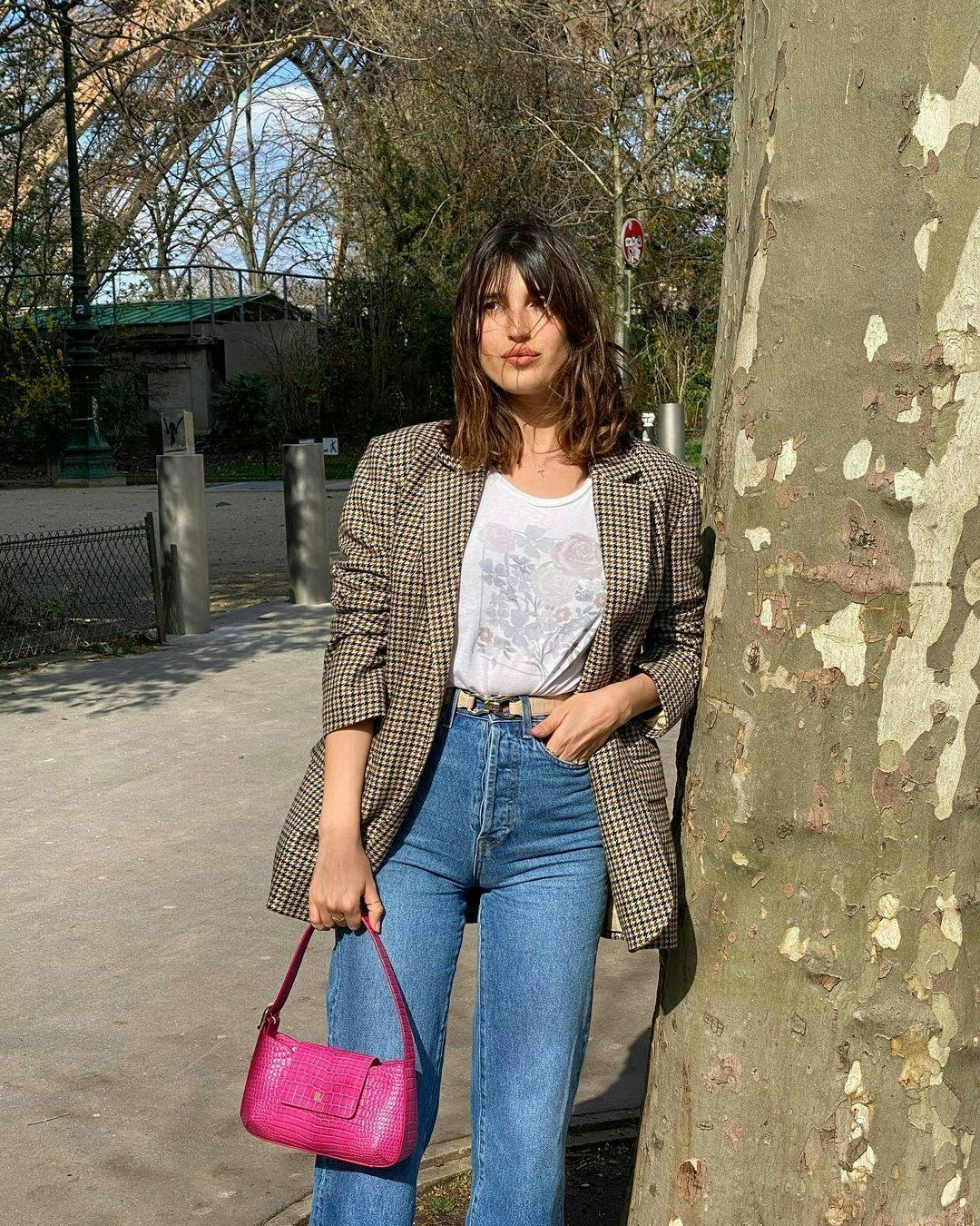 Jeanne Damas in jeans, a white t-shirt, a patterned blazer, and holds a bright pink bag.