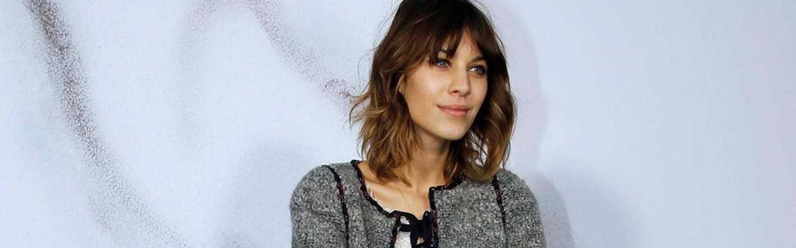 Alexa Chung wearing a tweed jacket, flowy white top, sheer black tights and black leather ankle boots and carrying a straw bag.
