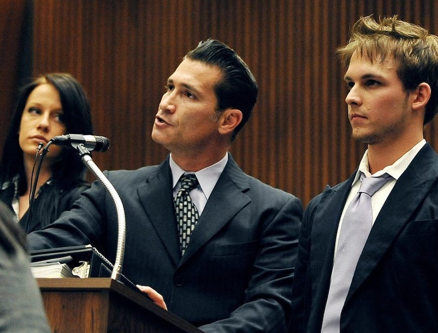 A lawyer speaking in the courtroom next to a man standing in a black suit.