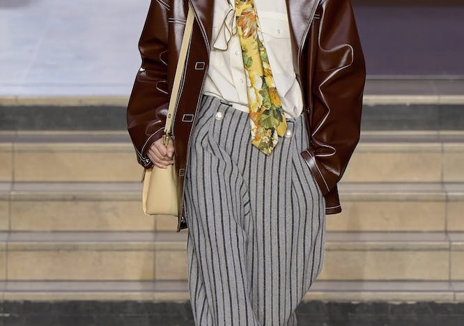 Model in a brown leather jacket, white blouse, and striped gray pants.