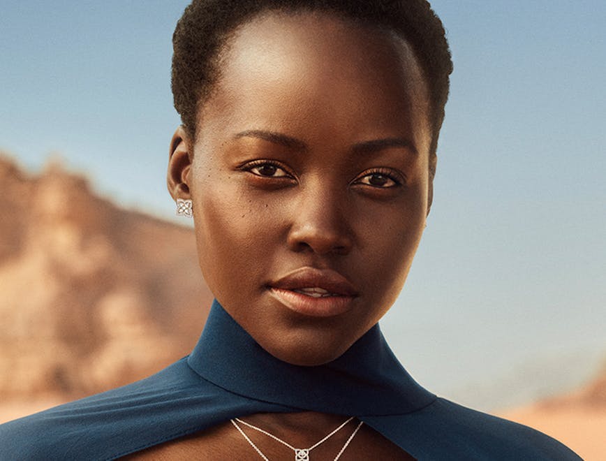image of lupita nyongo wearing a blue cape and dress wearing silver necklaces, rings, and a bracelet