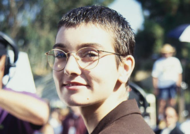young Sinéad O'Connor in glasses and a brown jacket looking at the camera.