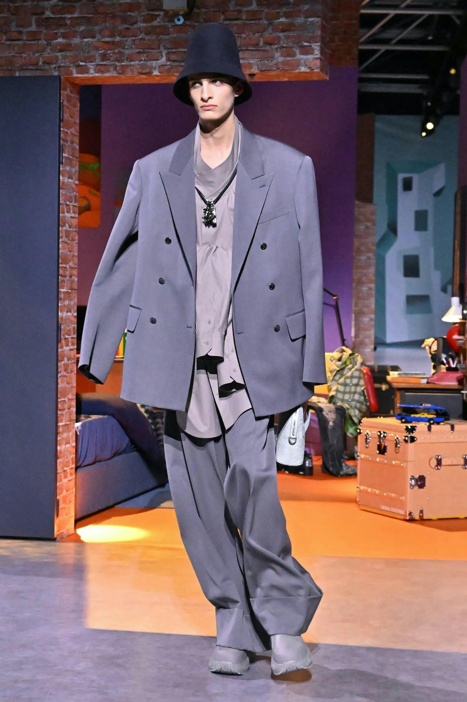 A model in a grey blazer, light purple blouse, and loose grey pants.