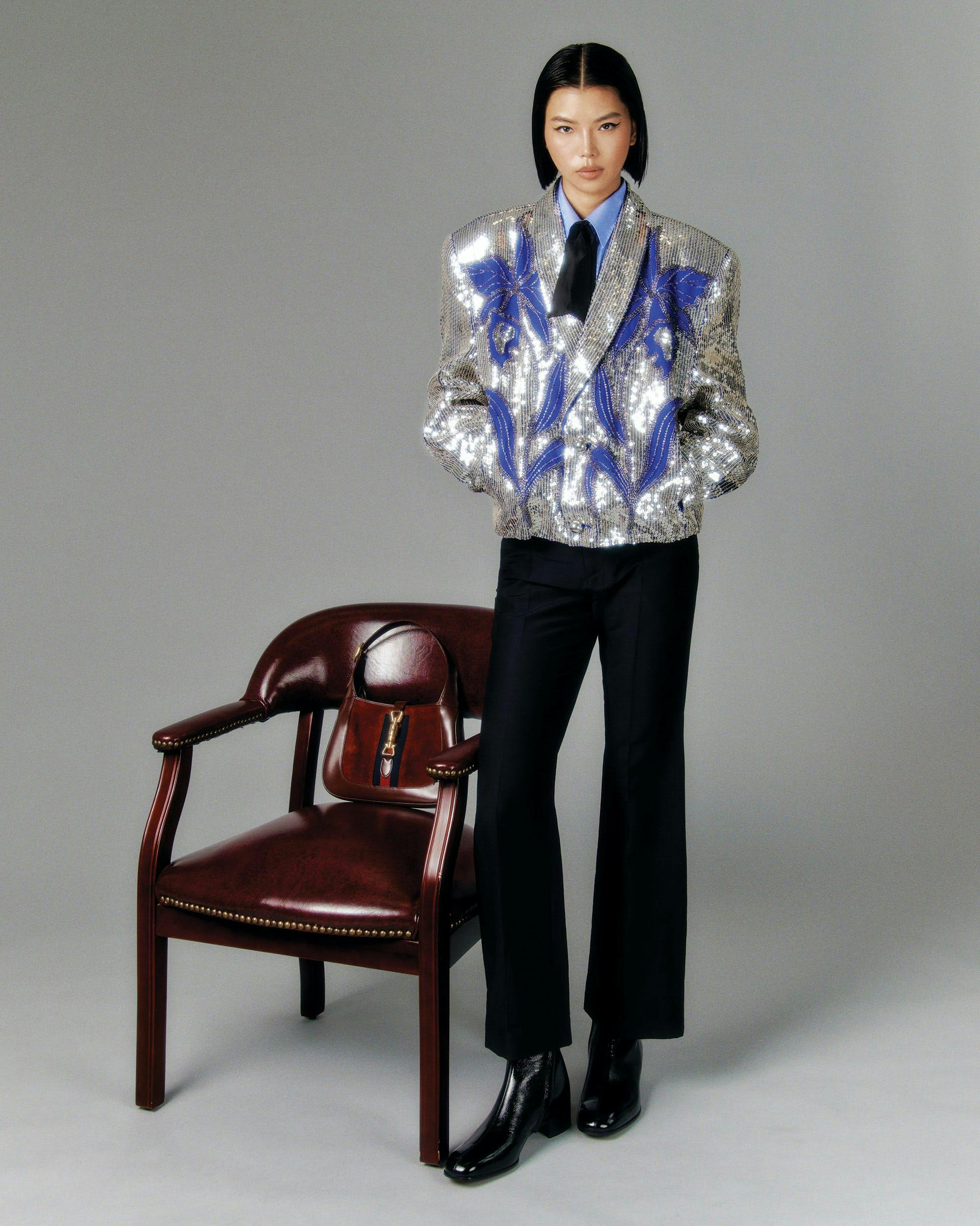 A model in a silver and blue sequined blazer and black pants next to a brown chair.