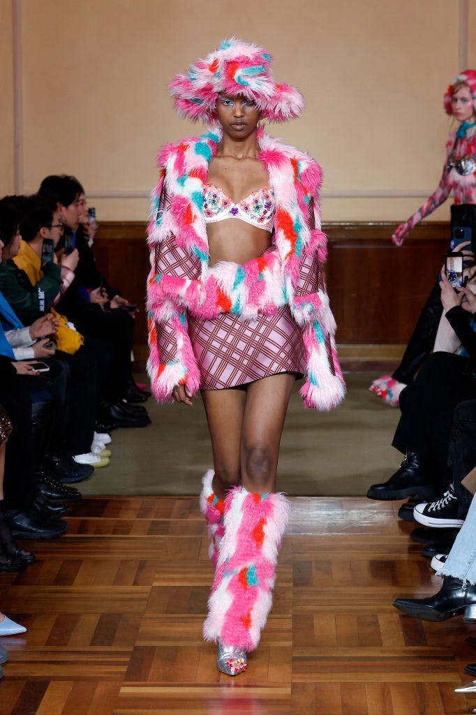 A model in a furry pink jacket, bra top, mini skirt, and boots.