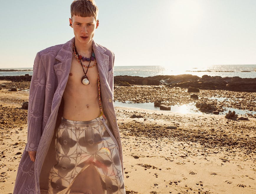 man on the beach wearing purple coat, white and gray printed pants, and multiple necklaces