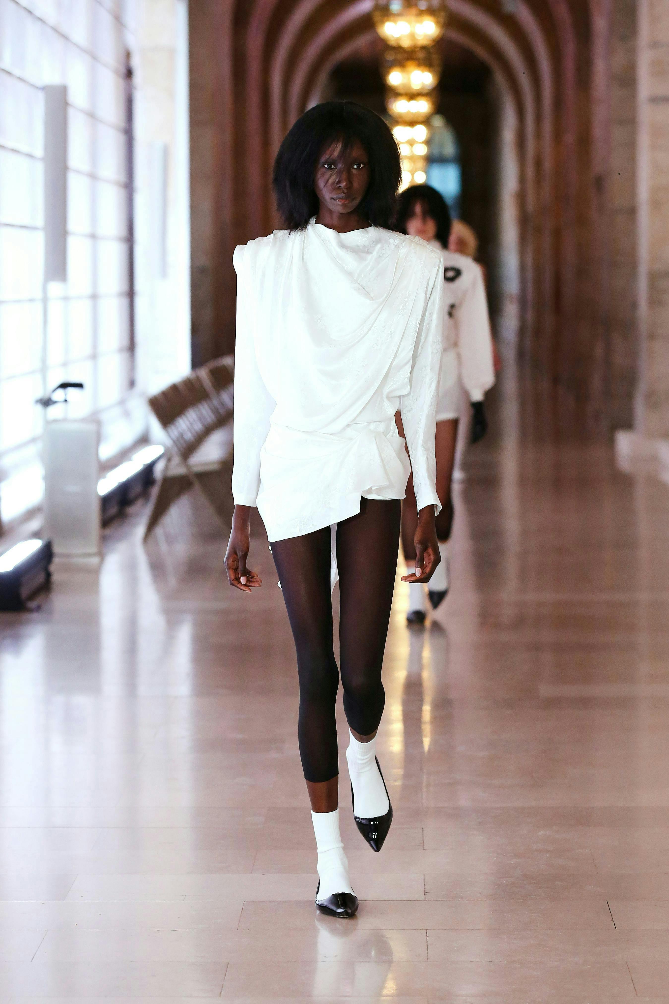 A model in a white blouse and shorts, black tights, white socks, and black flats.