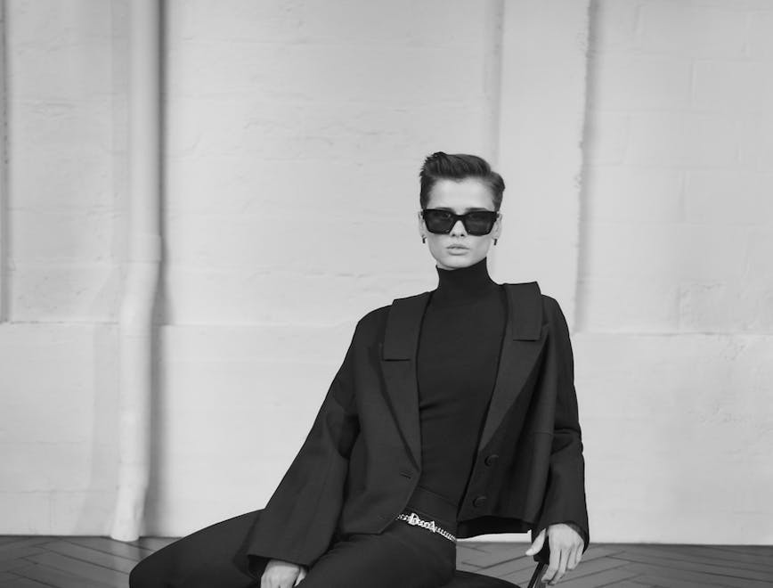 Black and white image of a model in all black with sunglasses sitting on a chair